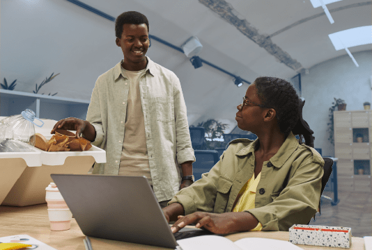 Green workplace. A young employee is speaking about a recycling program with an older employee sitting at a desk in front of a laptop