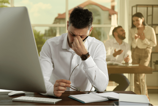 An employee is sitting at their desk, their glasses are in one hand, on top of the desk,. Their other hand is pinching the bridge of their nose. They look frustrated.