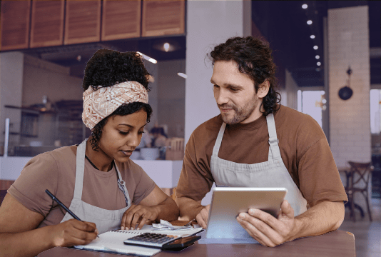 Two employees are standing at a high-top table, one is looking at a tablet, the other is using a paper, pencil, and calculator. They are goin over how to calculate overtime.