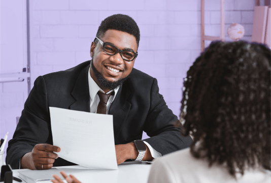 The Best Interview Questions to Ask: An interviewer is facing the camera and holding a resume. The interviewer is asking an interviewee questions across a desk. They are both facing each other and the interviewer is facing the camera.