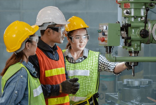 Hiring Co-op students: two young trainees are being shown a piece of industrial equipment by a senior employee. They are all wearing PPE like hard hats, safety glasses, gloves, and high-visibility vests.