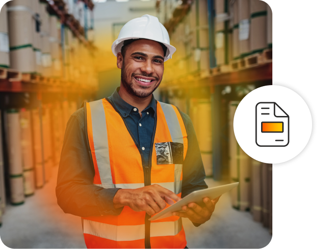 A man smiling wearing a hard helmet and high vis vest, holding a tablet in a warehouse.