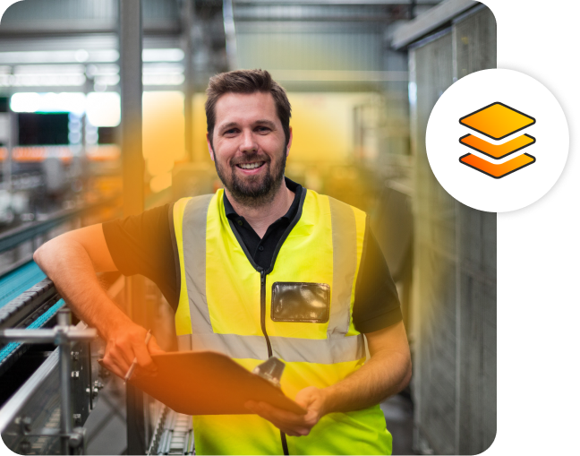 A man wearing a high vis vest, smiling at the camera while holding a clipboard in a factory.