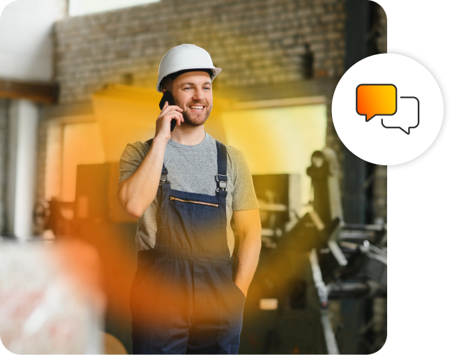 A man smiling wearing overalls and a hard helmet, while on a phone call in a workshop.