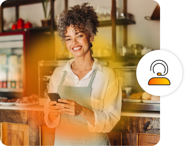 A woman smiling wearing an apron, holding a cell phone in a bakery.