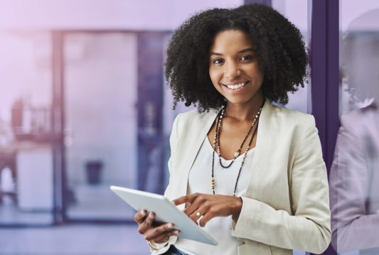 A professionally dressed woman is smiling while looking up from a tablet.