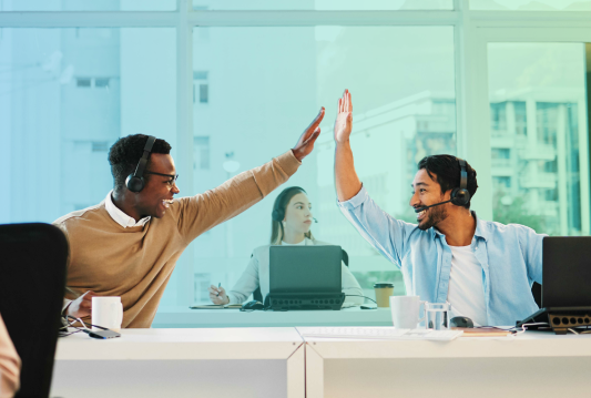 Two employees are sitting at their desks beside each other. They are happy and high-fiving each other.