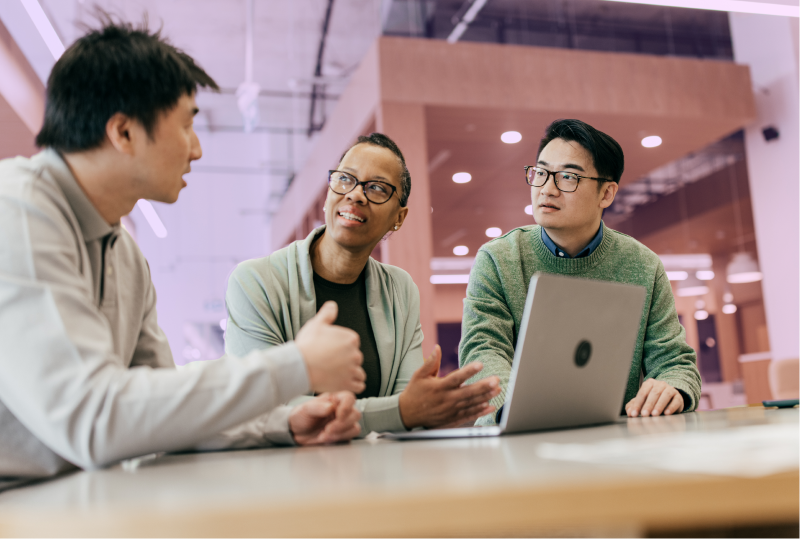 Three employees are in a shared workspace, and having a discussion around a laptop while using Canadian HR solutions