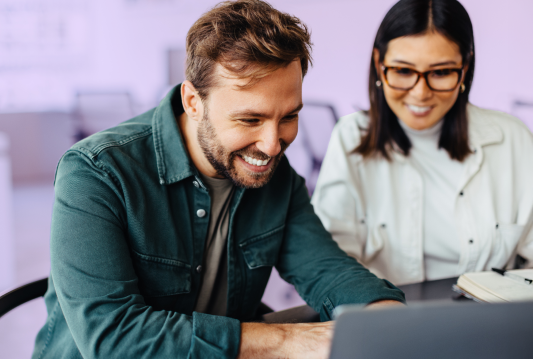 Two employees are seated at a desk and looking at a laptop. They are both smiling